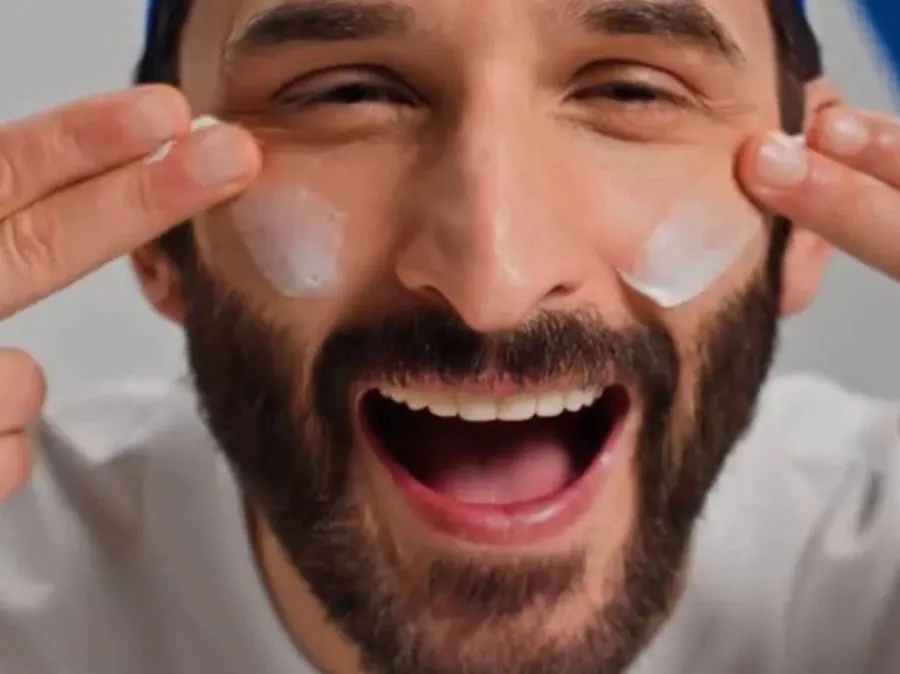 Homem usa o creme hidratante de CeraVe nas bochechas enquanto sorri para a câmera.